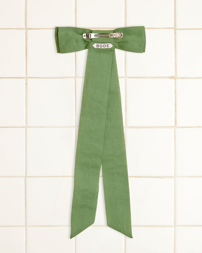 The Bow Barrette - Green, crafted from cotton voile with long tails, is elegantly hanging on a white tiled wall.