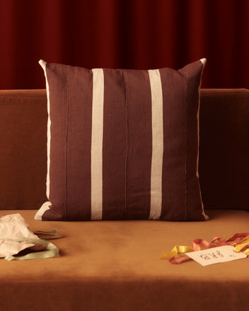 A Chimney Flue Pillow with brown and white stripes sits on a brown sofa, surrounded by papers and ribbons, blending perfectly with other home goods.