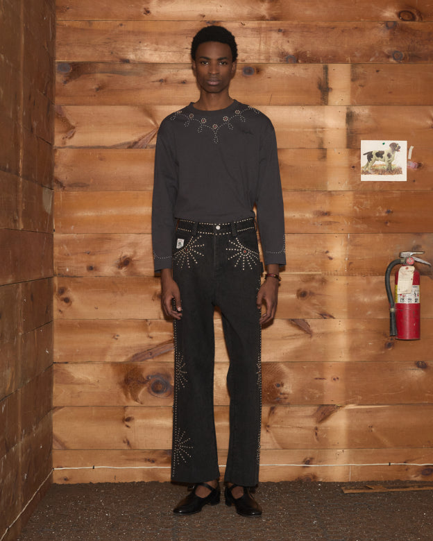 A person in the Silver Stud Long Sleeve Tee and black gemstone embellished pants stands in front of a wooden wall with a fire extinguisher.