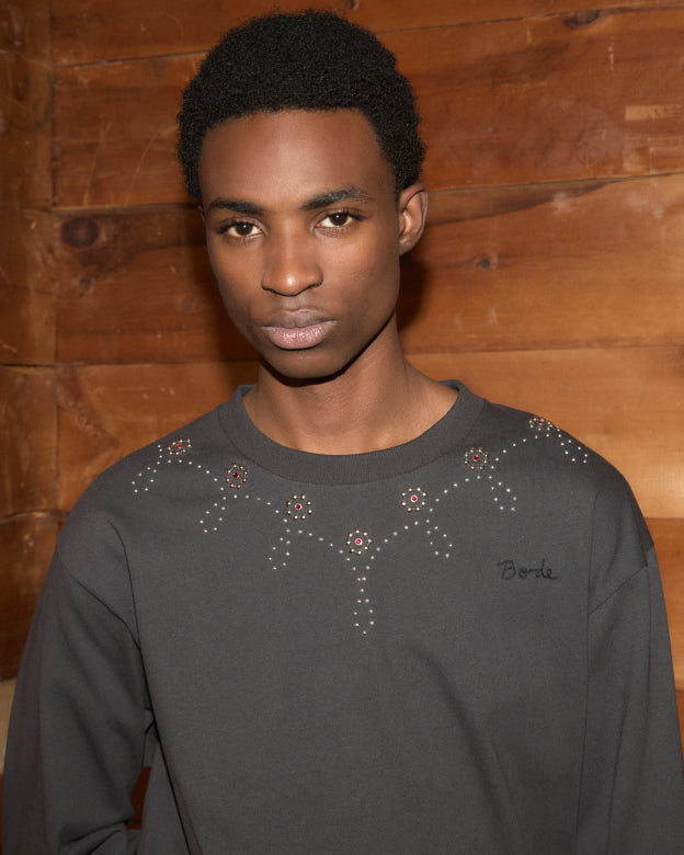 Wearing the Silver Stud Long Sleeve Tee with gemstone embellished designs, a person stands in front of a wooden wall, looking at the camera.