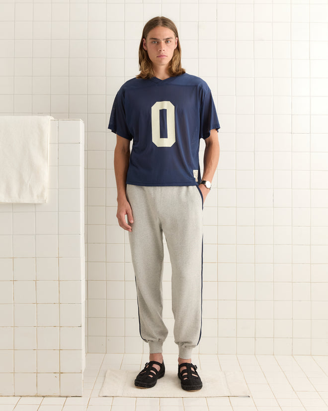A person with long hair, dressed in the Scrimmage Jersey - Navy featuring a "0" and gray sweatpants, stands in a tiled room.