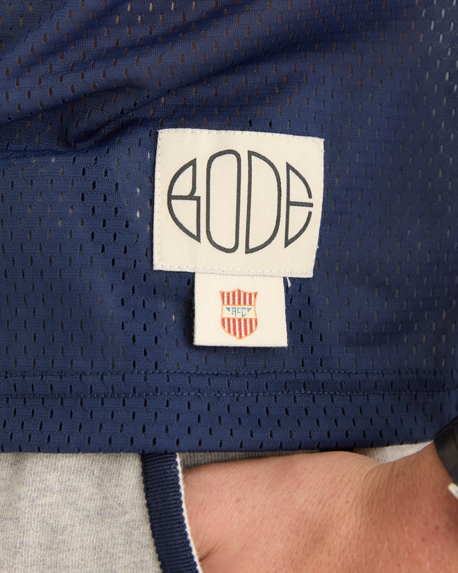 A close-up of the "BODE" label on navy blue mesh from the Scrimmage Jersey - Navy, featuring a small red and white shield tag, evoking a vintage football jersey style.