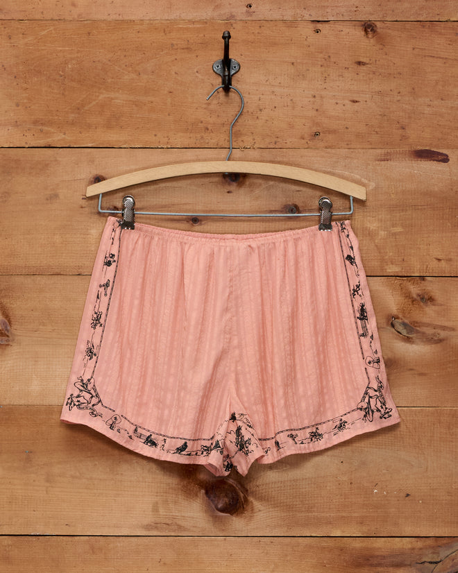 Bronco Buster Shorts in peach satin with black floral trim, inspired by jacquard silk tap shorts, displayed on a hanger against a wooden wall.
