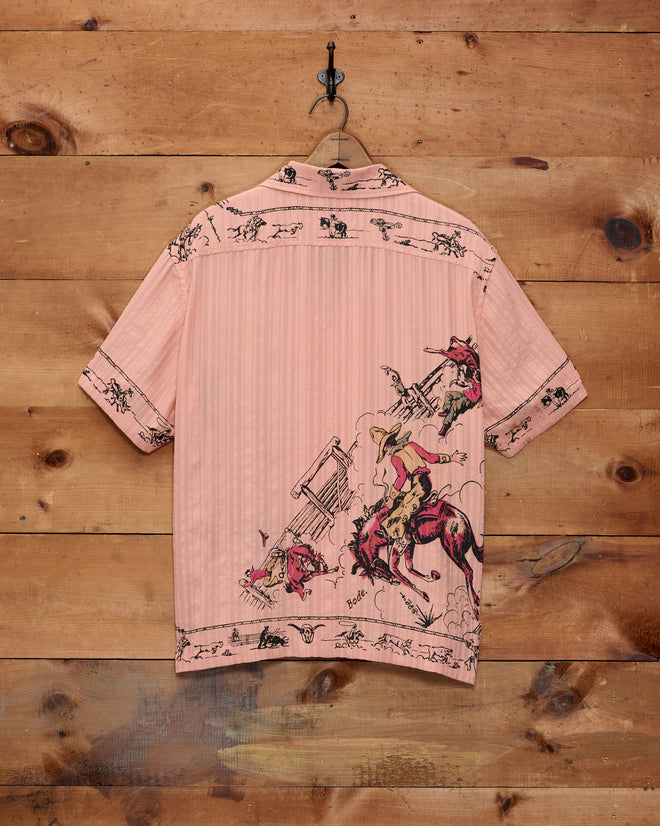 The Bronco Buster Shirt, a pink short-sleeve top featuring vibrant rodeo and horse illustrations, hangs on a hook against a wooden wall, perfectly capturing the spirit of a Cheyenne, Wyoming souvenir.