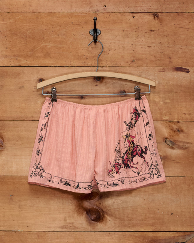 The Bronco Buster Shorts are pink jacquard silk tap shorts featuring a cowboy and horse print, inspired by a 1950s souvenir scarf from Cheyenne, Wyoming. Displayed on a wooden hanger against a wooden wall, these unique shorts evoke vintage Western charm.