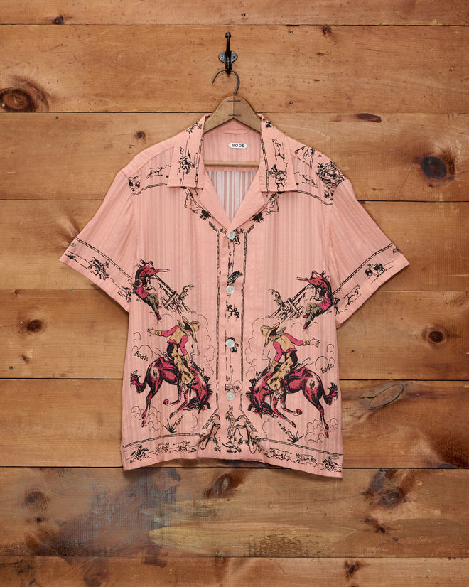 The Bronco Buster Shirt—a pink short-sleeve button-up featuring a playful horse design and cowboy print—hangs against a wooden wall, channeling the distinctive charm of Cheyenne, Wyoming souvenir style.