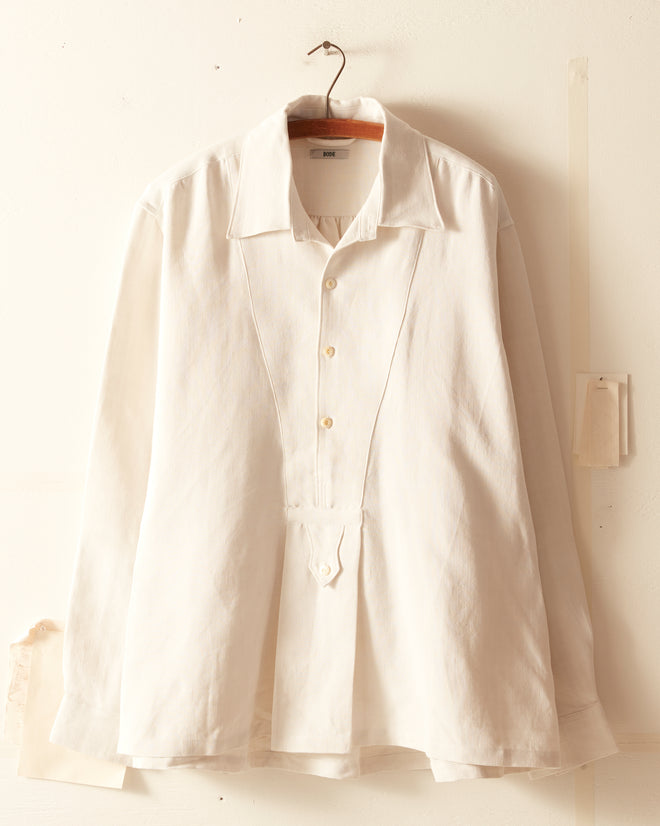 The Antique Linen Manchester Shirt, a white long-sleeve men's smock with a pleated front, hangs on a thin metal hanger against a white wall.