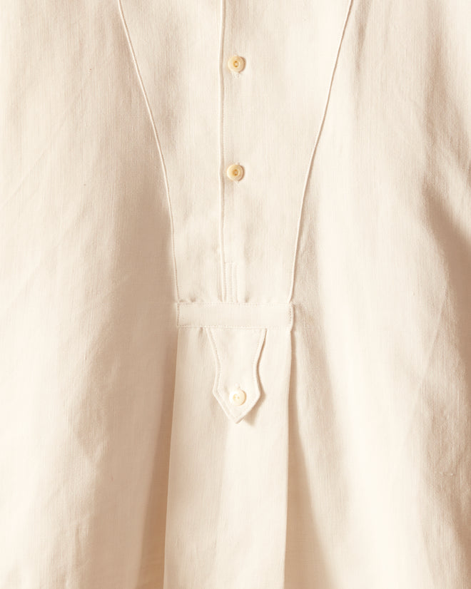 The Antique Linen Manchester Shirt, a white long-sleeve men's smock with a pleated front, hangs on a thin metal hanger against a white wall.