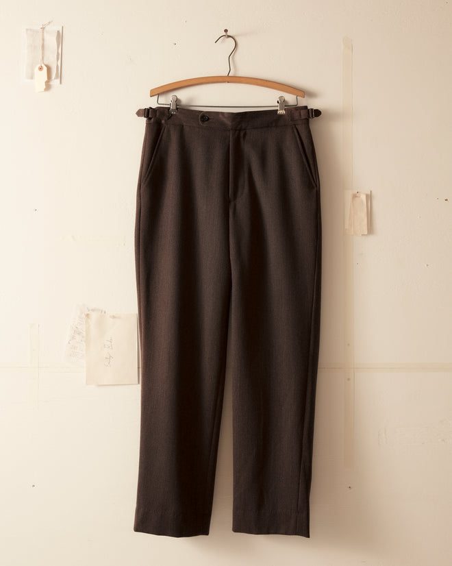 Twill Side Buckle Trousers in black, crafted from 100% wool, hang elegantly on a wooden hanger against a light wall, echoing timeless midcentury suiting.