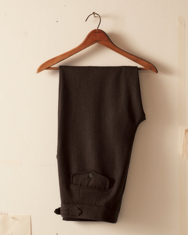 The Twill Side Buckle Trousers in black, made of 100% wool, are neatly folded over a wooden hanger against a light-colored wall.