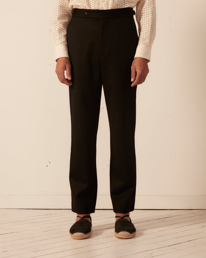 A person stands on a wooden floor wearing sleek black Twill Side Buckle Trousers paired with a white textured shirt.