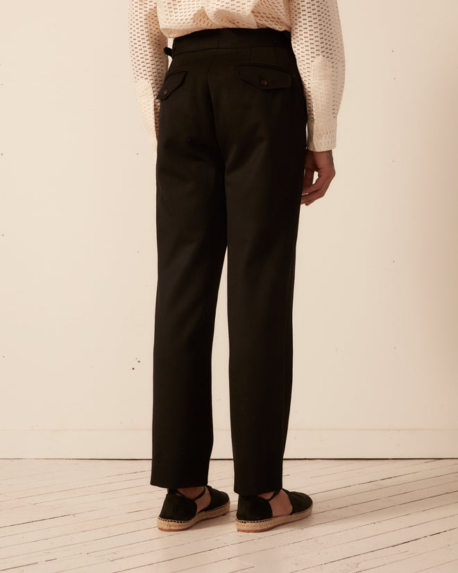 Person in stylish Twill Side Buckle Trousers - Black and a textured white top, standing on a light wooden floor, facing away from the camera.