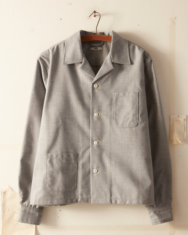 The Burford Overshirt, a gray button-up shirt adorned with antique pearl buttons, hangs on a wooden hanger against a white wall. Crafted from deadstock wool, this shirt features a collar, long sleeves, and three pockets on the front. This one-of-a-kind piece adds timeless elegance to any wardrobe.