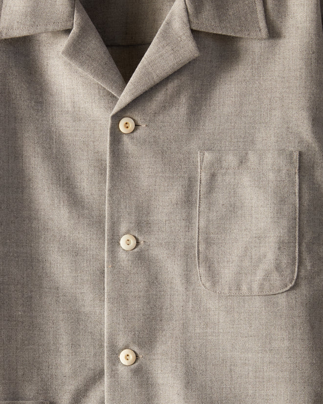 The Burford Overshirt, a gray button-up shirt adorned with antique pearl buttons, hangs on a wooden hanger against a white wall. Crafted from deadstock wool, this shirt features a collar, long sleeves, and three pockets on the front. This one-of-a-kind piece adds timeless elegance to any wardrobe.