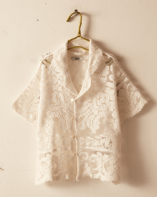 The Cloudy Lace Kids' Shirt, a charming white crocheted short-sleeve shirt featuring delicate filet lace detail, is perfect for children ages 2-6 and displayed on a hanger against a cream wall.