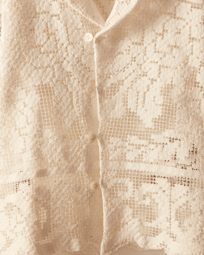 The Cloudy Lace Kids' Shirt, a charming white crocheted short-sleeve shirt featuring delicate filet lace detail, is perfect for children ages 2-6 and displayed on a hanger against a cream wall.