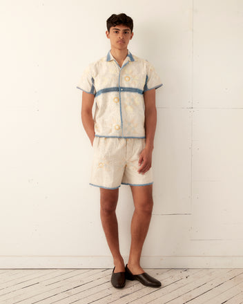 A person stands against a plain white wall wearing the Crochet Ladderwork Short Sleeve Shirt and matching shorts, both featuring blue trim details reminiscent of a vintage tablecloth shirt. They have short hair and are wearing dark loafers.