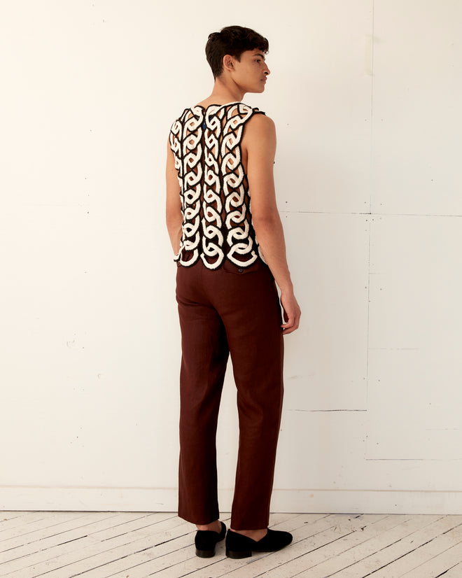 A person poses against a white wall, wearing the Crochet Ring Tank—a sleeveless top featuring an interwoven design reminiscent of a handmade crochet vest from the 1970s—paired with brown pants.