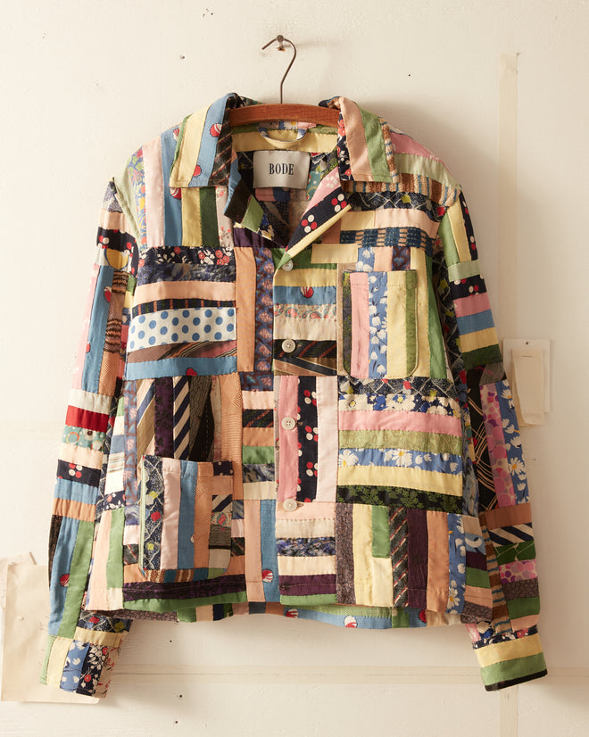 A vibrant Fiesta Stripe Shirt, available in size S/M, crafted from a variety of patterned fabric strips reminiscent of a vintage quilt shirt. It features a collared neck and button-down front and is displayed on a simple wire hanger against a plain wall.