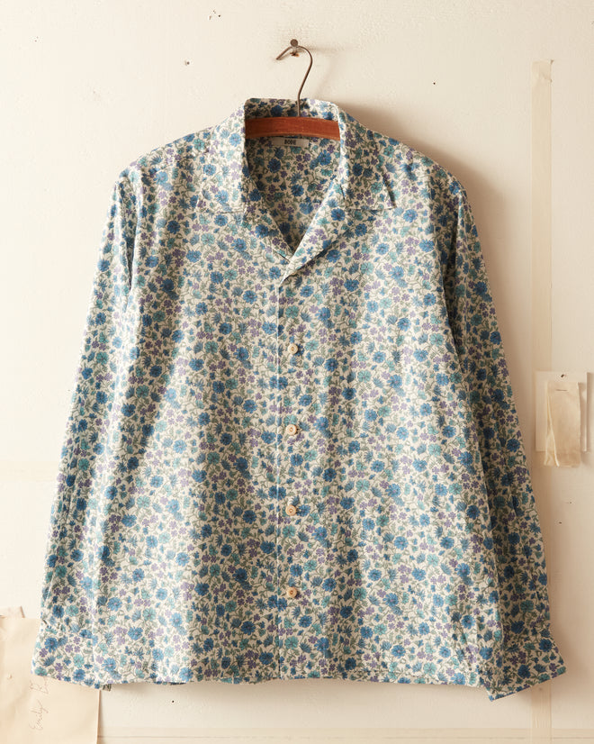 The Frozen Field Shirt, featuring a blue and green floral pattern and crafted from unique textile, hangs on a wooden hanger against a cream-colored wall.