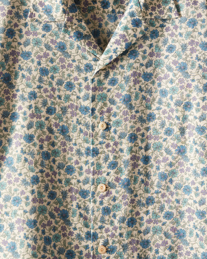 The Frozen Field Shirt, featuring a blue and green floral pattern and crafted from unique textile, hangs on a wooden hanger against a cream-colored wall.