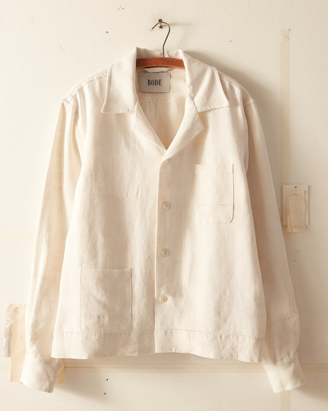 The Ladder Egg Shell Shirt, a Bode creation, features a white button-down design with a collar, front pockets, and intricate ladderwork embroidery. This unique piece hangs on a metal hook against a white wall and is crafted from antique French linens.