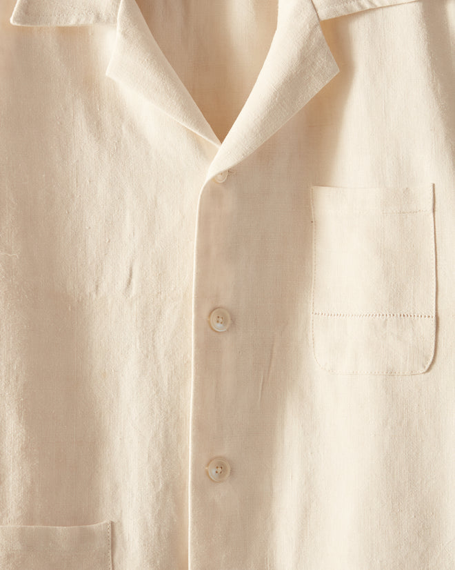 The Ladder Egg Shell Shirt, a Bode creation, features a white button-down design with a collar, front pockets, and intricate ladderwork embroidery. This unique piece hangs on a metal hook against a white wall and is crafted from antique French linens.