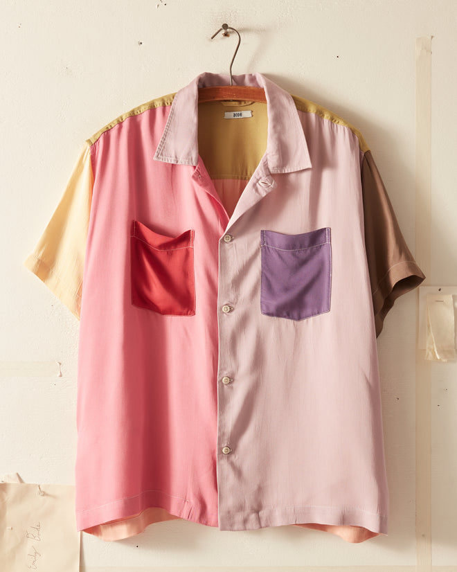 A Buckaround Short Sleeve Shirt with a vibrant color-block design hangs on a hook. The shirt, featuring multicolored buttons, boasts two front pockets in different hues: red and purple.