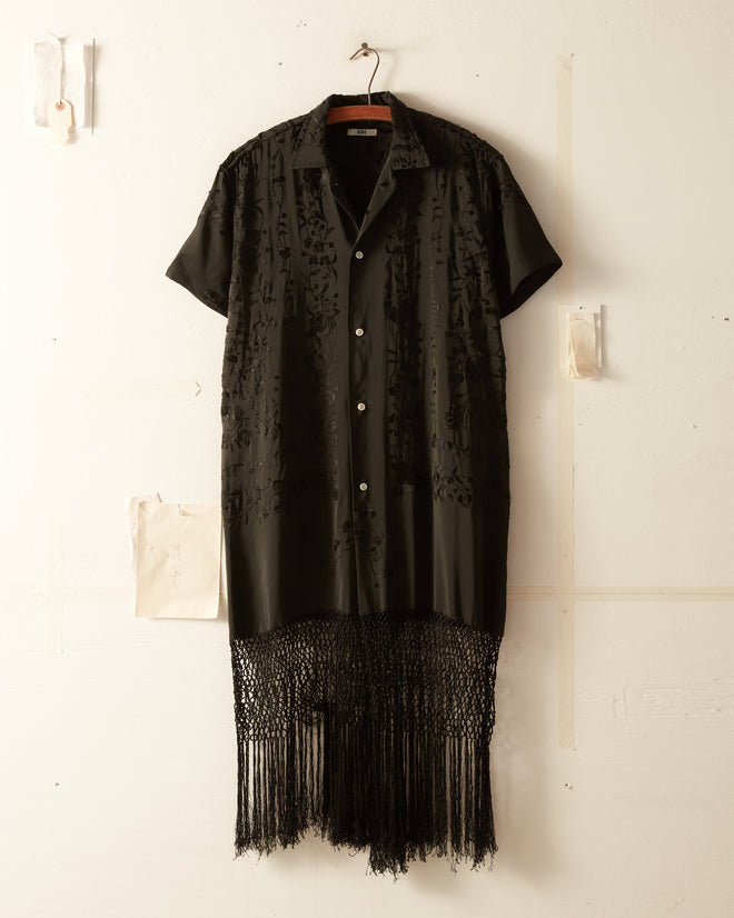 A Raven Floral Tunic - S/M, featuring intricate black embroidery on the chest and a fringed hemline reminiscent of unique vintage clothing, hangs against a white wall.