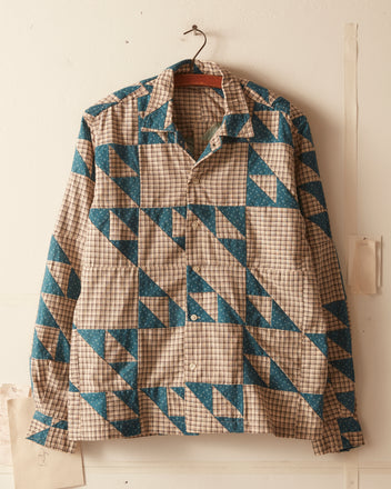 The Wandering Lover Quilt Long Sleeve Shirt - Blue, made from 100% cotton and showcasing a geometric pattern of blue and beige triangles and grids, hangs on a hanger against a light-colored wall.